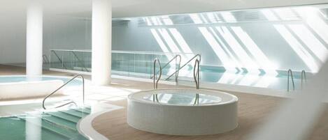 A modern indoor swimming pool with clear glass walls, a central circular hot tub, and a wooden floor.