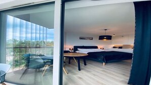 A modern hotel room with a large sliding glass door leading to a balcony, a bed with a black headboard, a bedside table with a lamp, and a small round table. - Apartment in Pobierowo near Beach & SPA (Pobierowo)