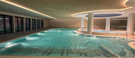 An indoor swimming pool with a tiled floor, a central hot tub, and a poolside area with a railing.