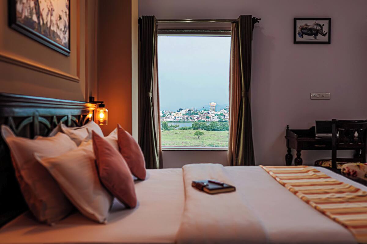 A hotel room with a bed, pillows, a bedside table, a lamp, and a view of a cityscape through a window.