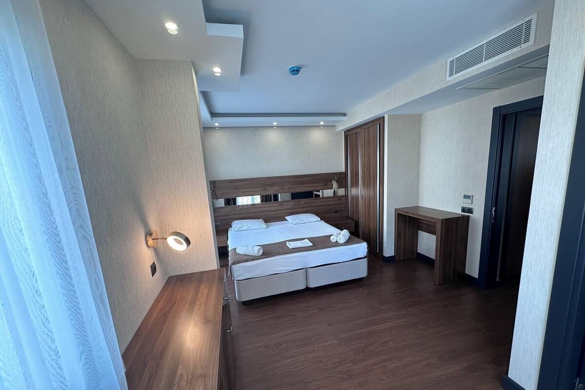 A modern hotel room with a wooden bed, a bedside table, a desk, and a wardrobe.