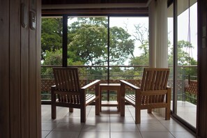 A balcony with two wooden chairs and a small table, large windows with a view of trees, and a wooden door. - Hotel Ficus Monteverde (Monteverde)