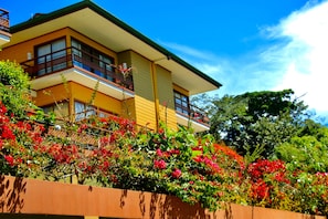 A multi-story building with balconies, surrounded by lush greenery and flowering plants.