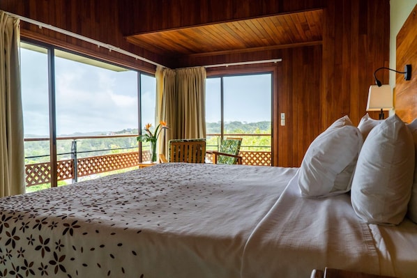 A hotel room with a large bed, wooden walls, a view through large windows, and a small table with a chair.