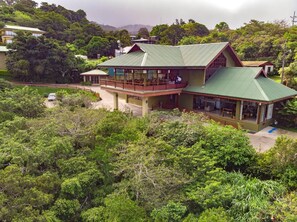 A two-story building with a green roof, surrounded by dense greenery and hills in the background.