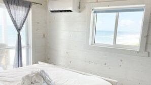 Een slaapkamer met een houten plafond, witte muren, een groot bed met witte lakens, een raam met uitzicht op het strand en een airconditioner.