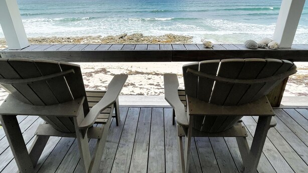 Een houten terras met twee Adirondack-stoelen uitkijkend over een strand met golven en een heldere lucht.