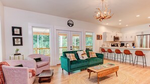 A spacious living area with a green sofa, a pink armchair, and a wooden coffee table. There is a kitchen area with white cabinets and a stainless steel refrigerator. - Retreat - Creek, Sliding Rock, Waterfalls, Fishing, Hiking, Hot Tub, Fire Pit (Old Fort)