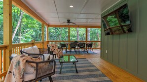 A covered outdoor area with wooden flooring, a ceiling fan, a table with chairs, and a TV mounted on the wall. - Retreat - Creek, Sliding Rock, Waterfalls, Fishing, Hiking, Hot Tub, Fire Pit (Old Fort)
