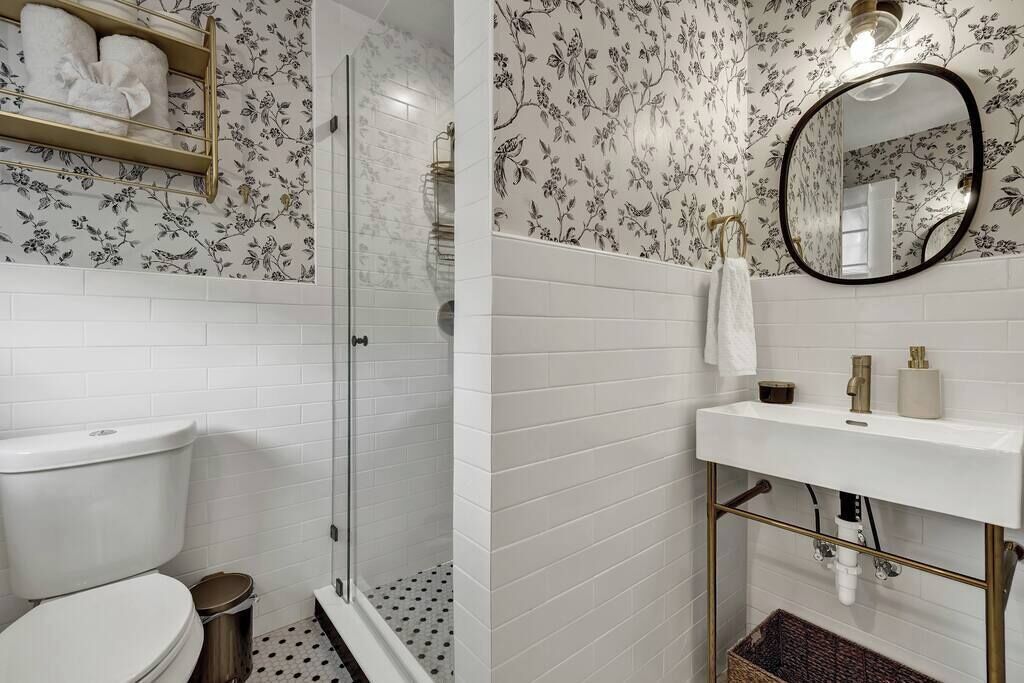 Bagno con pareti piastrellate di bianco, un WC, una doccia con porta in vetro, un lavandino con cornice dorata e uno specchio.