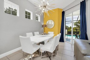A dining area with a white table and chairs, a round mirror on the wall, and a view of a swimming pool through large windows.