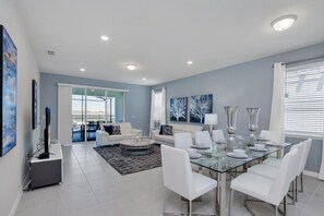 A modern living room with a glass dining table, white sofas, and a large window with blinds.