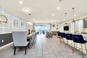 A modern kitchen with a dining area, featuring a long dining table with chairs, a kitchen counter, and a large window providing a view of the outdoors.
