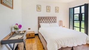 Ein Schlafzimmer mit einem Bett, einem Holztisch, einem Stuhl, einem kleinen Tisch mit Blumen und einem Fenster mit Fensterläden.