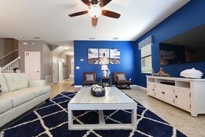 Ein Wohnzimmer mit einem weißen Sofa, einem Couchtisch und zwei Sesseln. Es gibt einen Deckenventilator, ein Bild an der Wand und einen Fernseher.