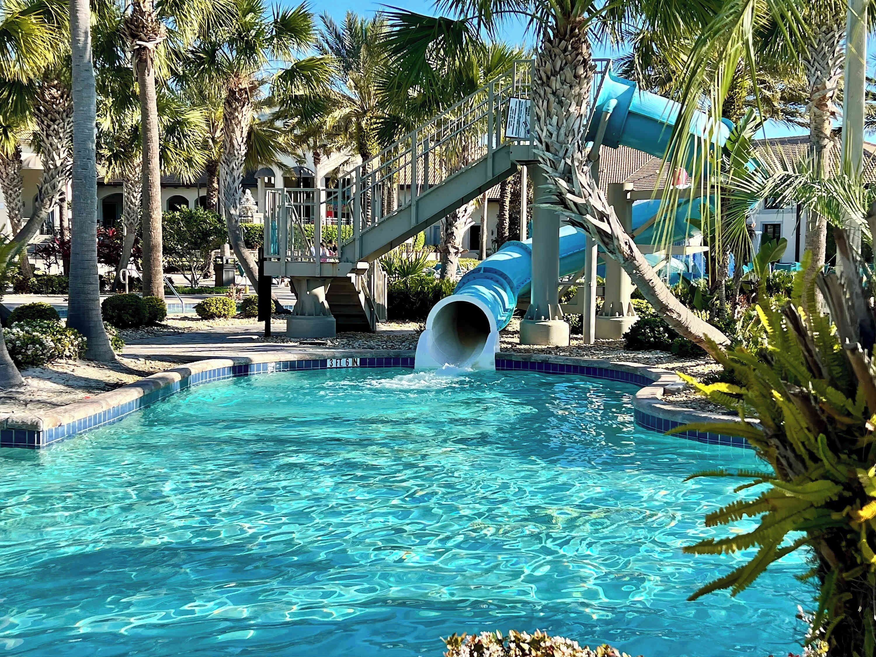 A swimming pool with a blue slide, surrounded by palm trees and a building with a balcony.