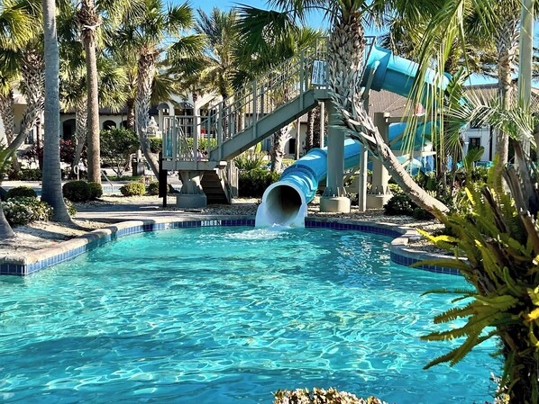 Une piscine avec un toboggan bleu, entourée de palmiers et bordée d’un bâtiment doté d’un balcon.