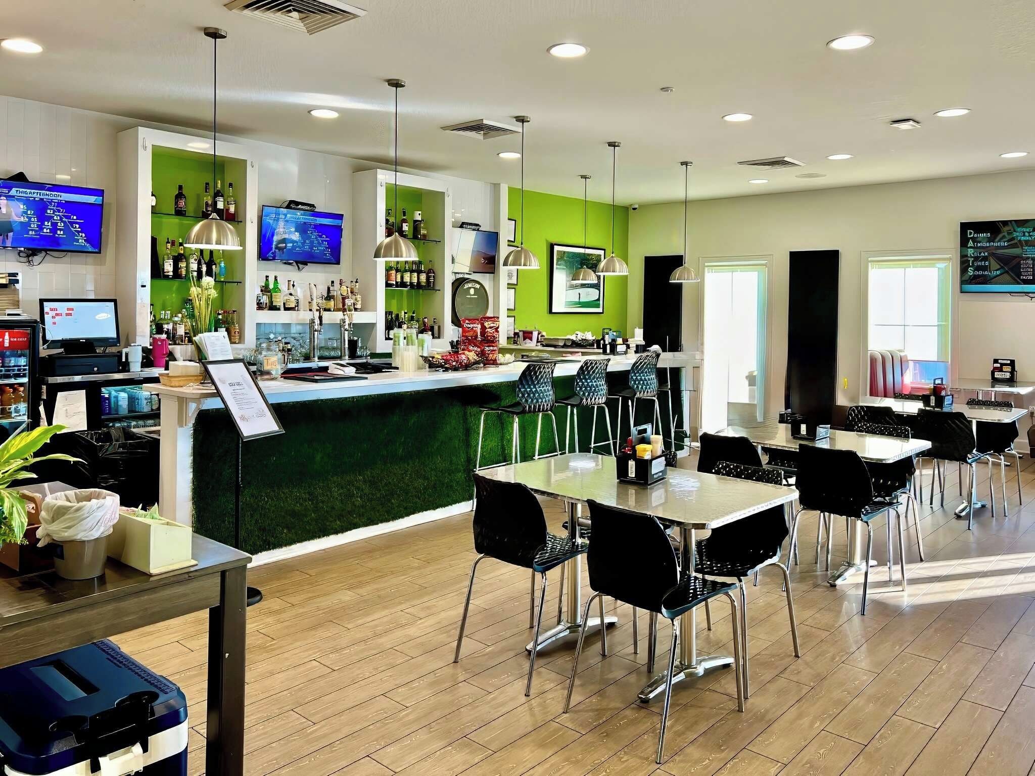Um espaço de bar moderno com uma parede verde decorativa, várias televisões e uma seleção variada de bebidas.