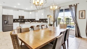 A modern kitchen with white cabinets, a stainless steel refrigerator, and a dining area with a wooden table and wicker chairs. - Private Pool & Spa | Game Room | Near Disney 8908 (Davenport)