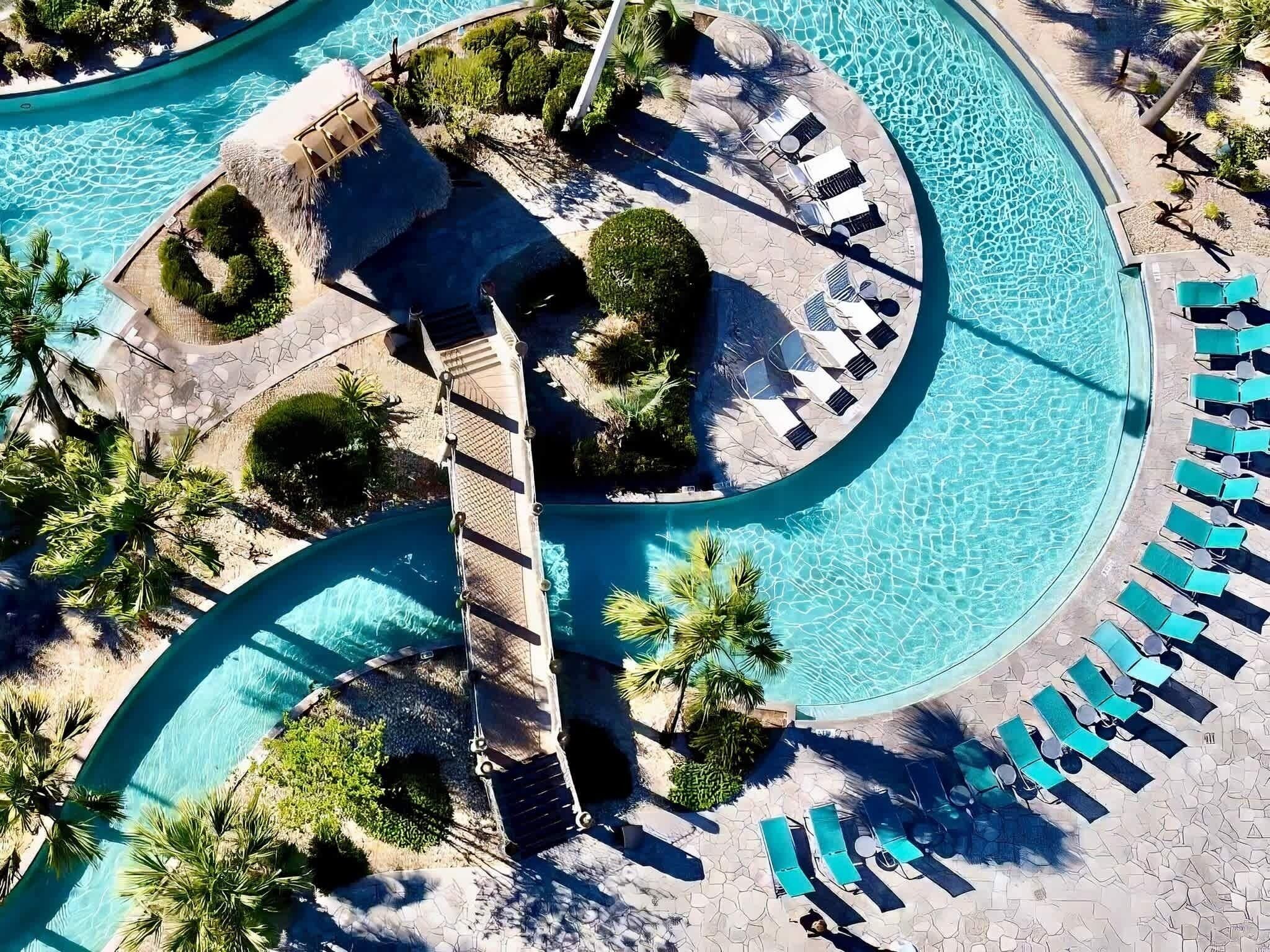 An aerial view of a resort with a large, winding pool, sun loungers, and palm trees.