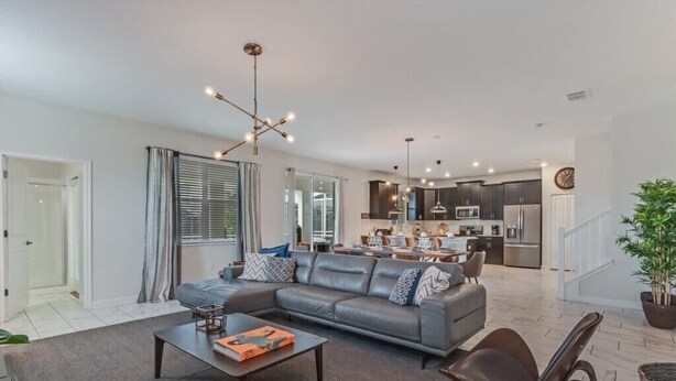 A modern living room with a grey sofa, coffee table, and chairs. A kitchen is visible in the background with dark cabinets and stainless steel appliances. - South Pool | Gameroom | Waterpark | Near Disney 8940 (Davenport)