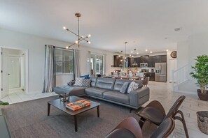 A modern living room with a grey sofa, coffee table, and chairs. A kitchen is visible in the background with dark cabinets and stainless steel appliances. - South Pool | Gameroom | Waterpark | Near Disney 8940 (Davenport)