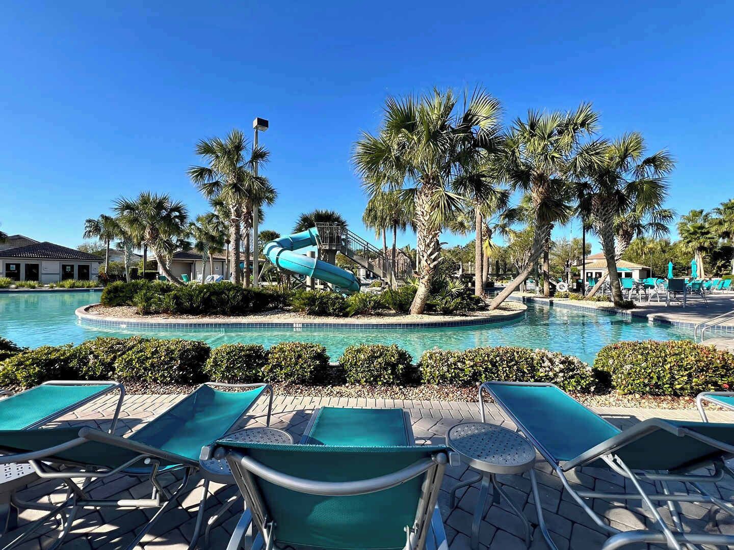 A pool area with a water slide, lounge chairs, palm trees, and a clear blue sky.
