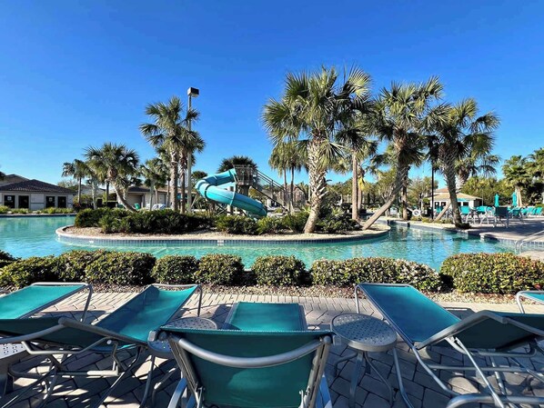 A pool area with a water slide, lounge chairs, palm trees, and a clear blue sky.