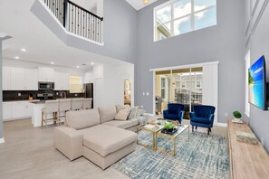 A spacious living area with a beige sectional sofa, a coffee table, and a blue armchair. A kitchen with white cabinets and a dining area with a blue chair are visible in the background.