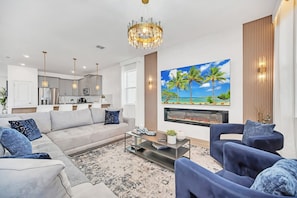 A modern living room with a large flat-screen TV, a fireplace, and a dining area with a kitchen in the background.