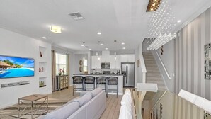Ein modernes Wohnzimmer mit einem großen Flachbildfernseher, einem grauen Sofa, einem Essbereich mit Stühlen, einer Küche mit weißen Schränken und einer Treppe.