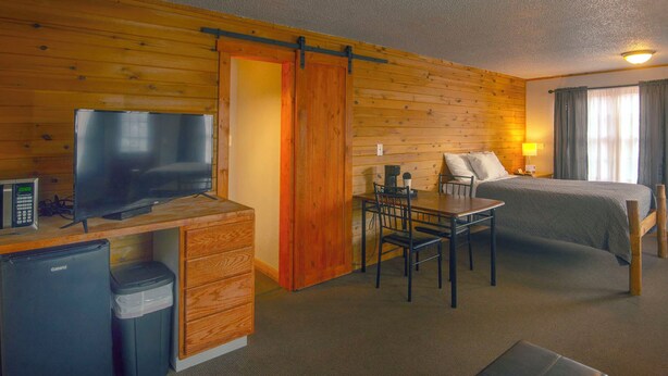 A compact hotel room with a wooden interior, a bed, a desk with a television, a small refrigerator, and a dining table. - Rustic Inn (Park Rapids)