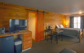 A compact hotel room with a wooden interior, a bed, a desk with a television, a small refrigerator, and a dining table. - Rustic Inn (Park Rapids)