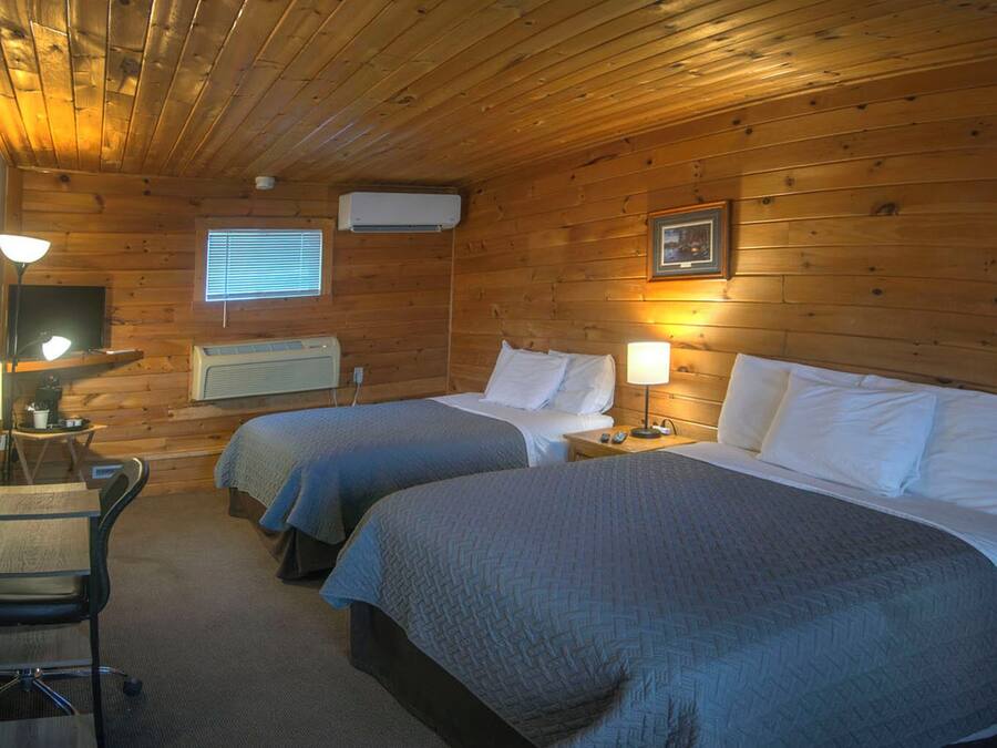 A cabin-style room with two beds, a desk with a chair, a television, and a window with blinds.