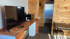 A compact kitchenette with a TV, coffee maker, and microwave. There is a small fridge, a wooden table, and a chair. A bathroom is visible through an open door. - Rustic Inn (Park Rapids)