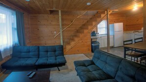 A log cabin interior with a living area featuring two dark-colored sofas, a coffee table, and a staircase leading to a kitchen area. - Rustic Inn (Park Rapids)
