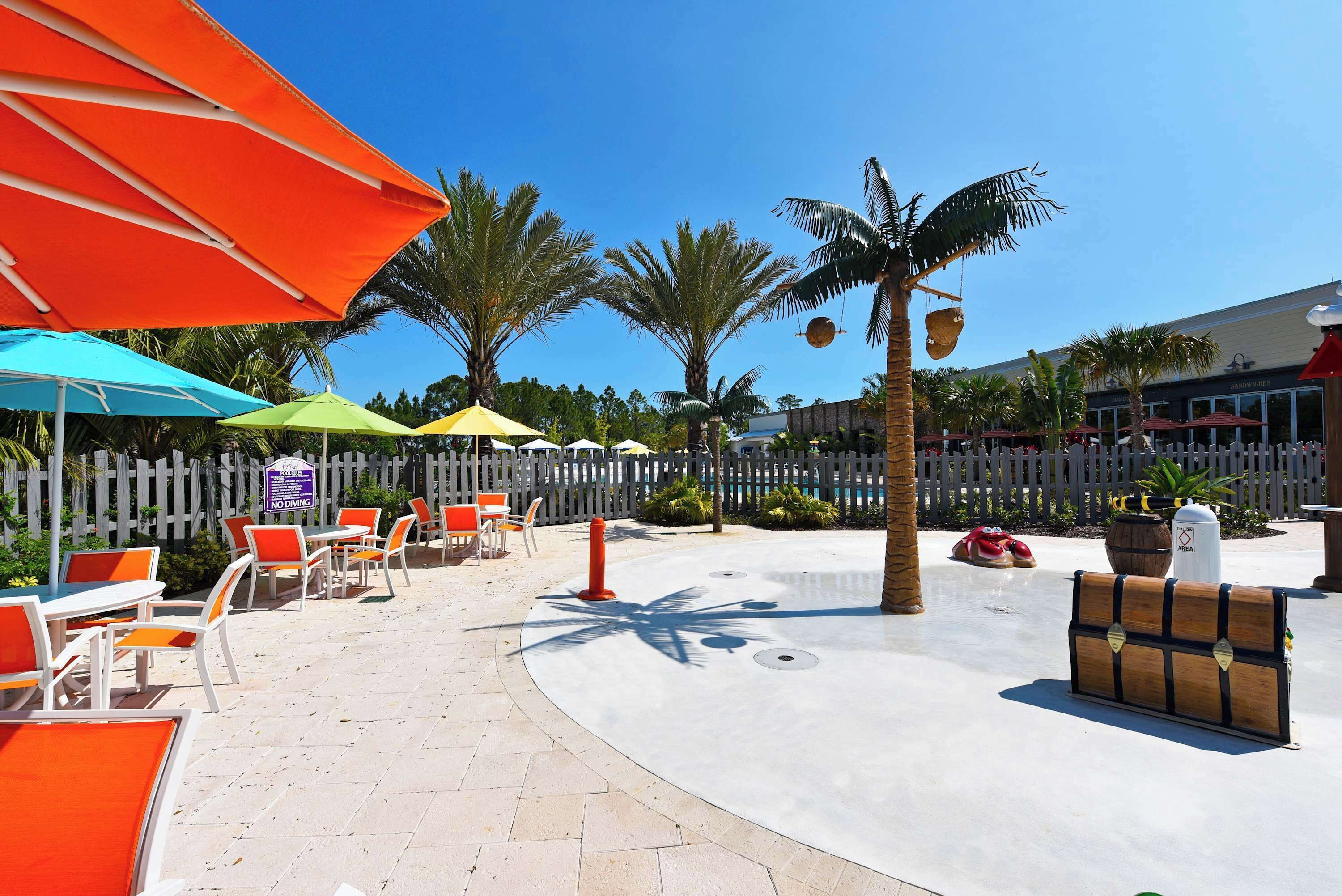 Zona junto a la piscina con mobiliario exterior, sombrillas y palmeras.