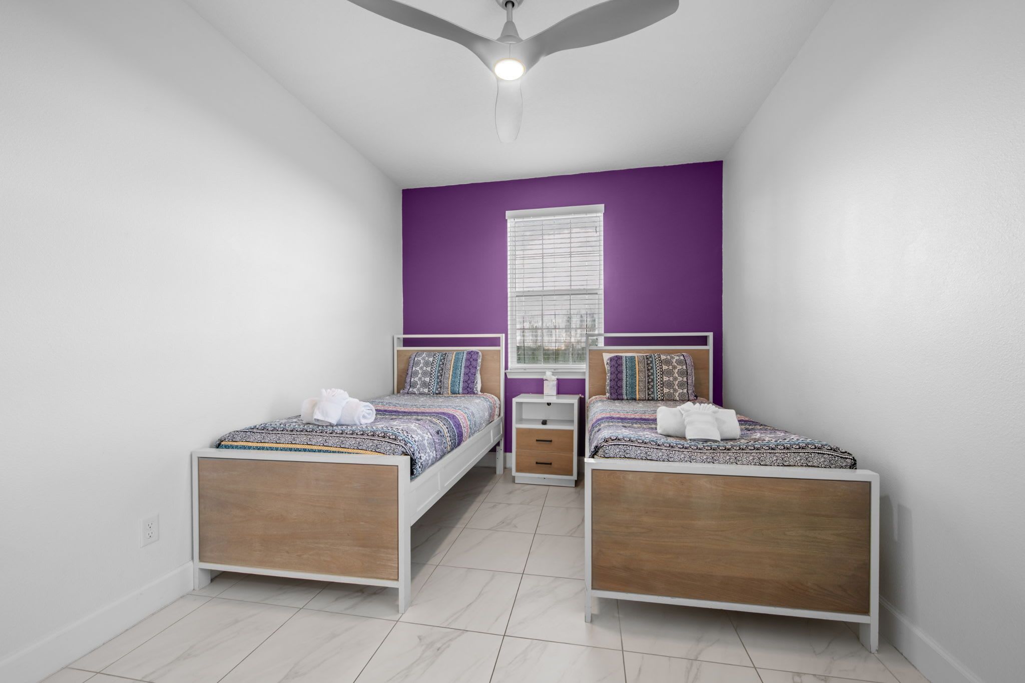 A bedroom with two beds, a ceiling fan, a window with blinds, and a white floor.