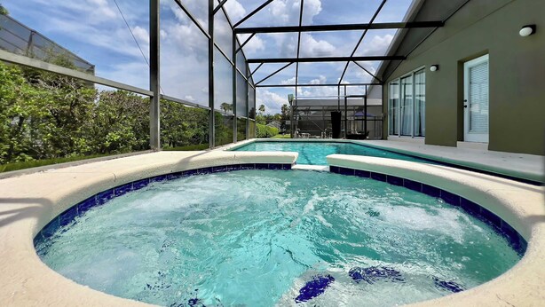 A covered outdoor swimming pool with a glass ceiling, surrounded by greenery and a house in the background. - Value | Private Pool | Game Room | Disney 1616 (Clermont)