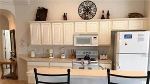 A kitchen with white cabinets, a microwave, a stove, and a refrigerator. There are two chairs with woven seats. - Value | Private Pool | Game Room | Disney 1616 (Clermont)