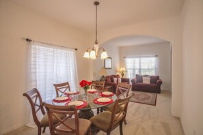 A dining area with a round table set for four, surrounded by wooden chairs. There is a chandelier above the table, and a sofa and lamp in the background.