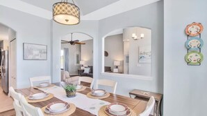 Un comedor con una mesa de madera dispuesta para cuatro personas, rodeada de sillas blancas. Hay un ventilador de techo, una lámpara fijada a la pared y un estante decorativo para platos en la pared.