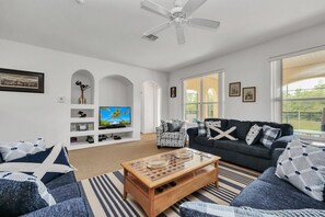 A modern living room with a flat-screen TV, a ceiling fan, and a large window providing a view of the outdoors. - Conservation View | Pool & Spa | 7 Miles Disney 203 (Davenport)