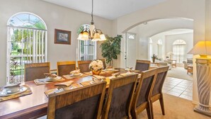 A dining area with a wooden table set for six, surrounded by chairs. There is a chandelier above the table, and a potted plant is visible to the side. - Conservation View | Pool & Spa | 7 Miles Disney 203 (Davenport)