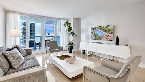 A modern living room with a sofa, a coffee table, a TV, and a large window with a city view. - Sauna | Hot Tub | Skyline & Bay Views | StayCozy (Miami)
