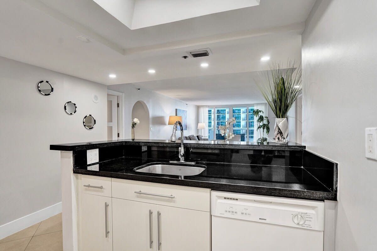 A modern kitchen with a black countertop, white cabinets, and a dishwasher. There are decorative mirrors on the wall and a plant in the corner.