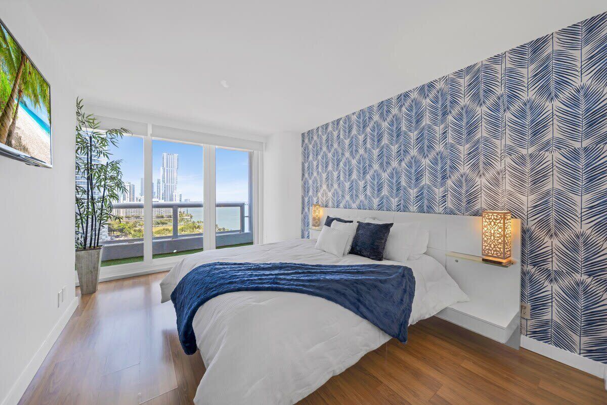 A bedroom with a large window, a bed, a nightstand, and a patterned wall.