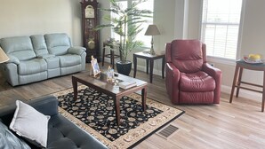 A living room with a green sofa, a red armchair, a coffee table, a potted plant, a clock, and a window with blinds.