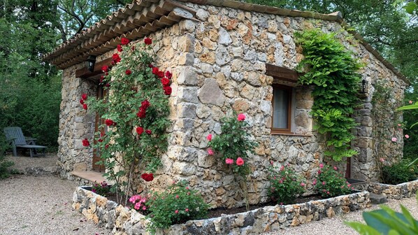 A stone cottage with a wooden roof, surrounded by a gravel path and greenery.
