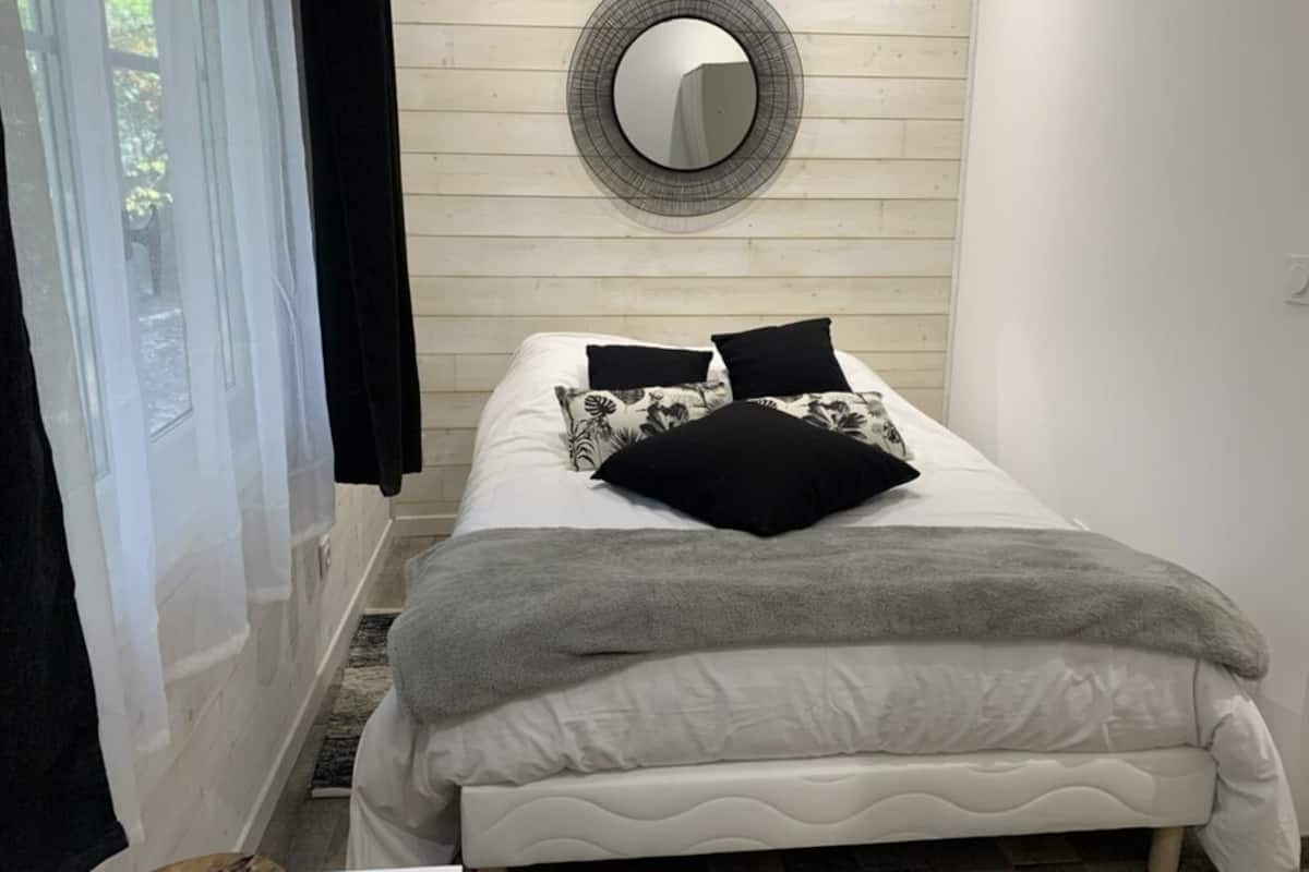 Una cama con cabecera blanca, manta gris y almohadas con estampado blanco y negro. Un espejo redondo cuelga de la pared sobre la cama.
